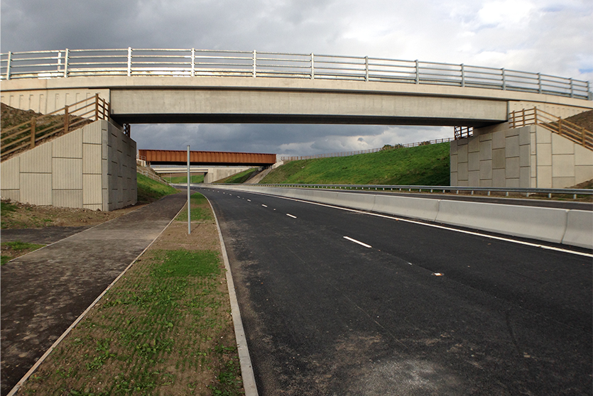 A construction view of the A6 Manchester Airport Relief Road project, showcasing a large retaining wall built using Geoquest Co. Ltd.'s precast concrete facing panels. The image highlights the integration of the road with surrounding infrastructure, illustrating the scale of the project and the innovative design solutions employed for the retaining walls and bridge abutments.